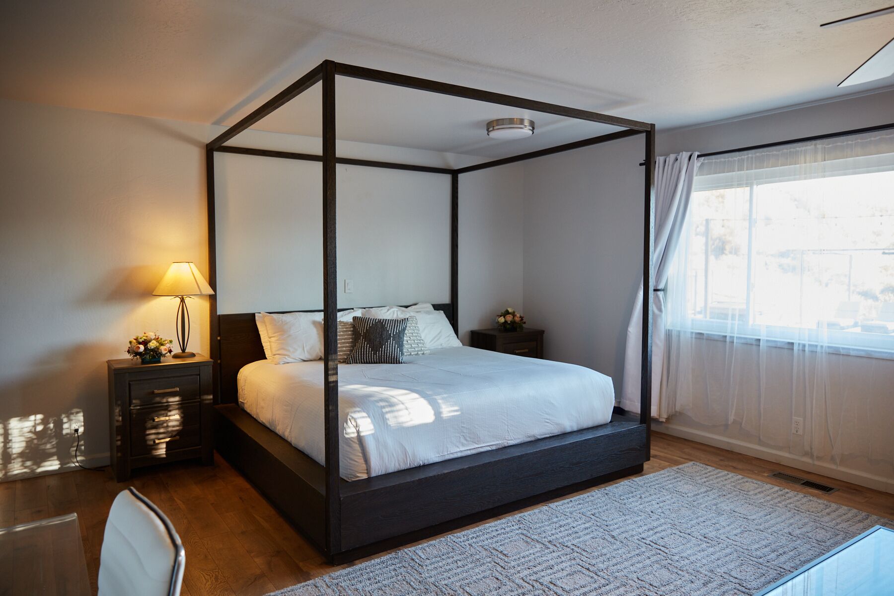A four-poster bed with a dark frame, a bedside table with a lamp, and a view of the outdoors through a window with sheer curtains.