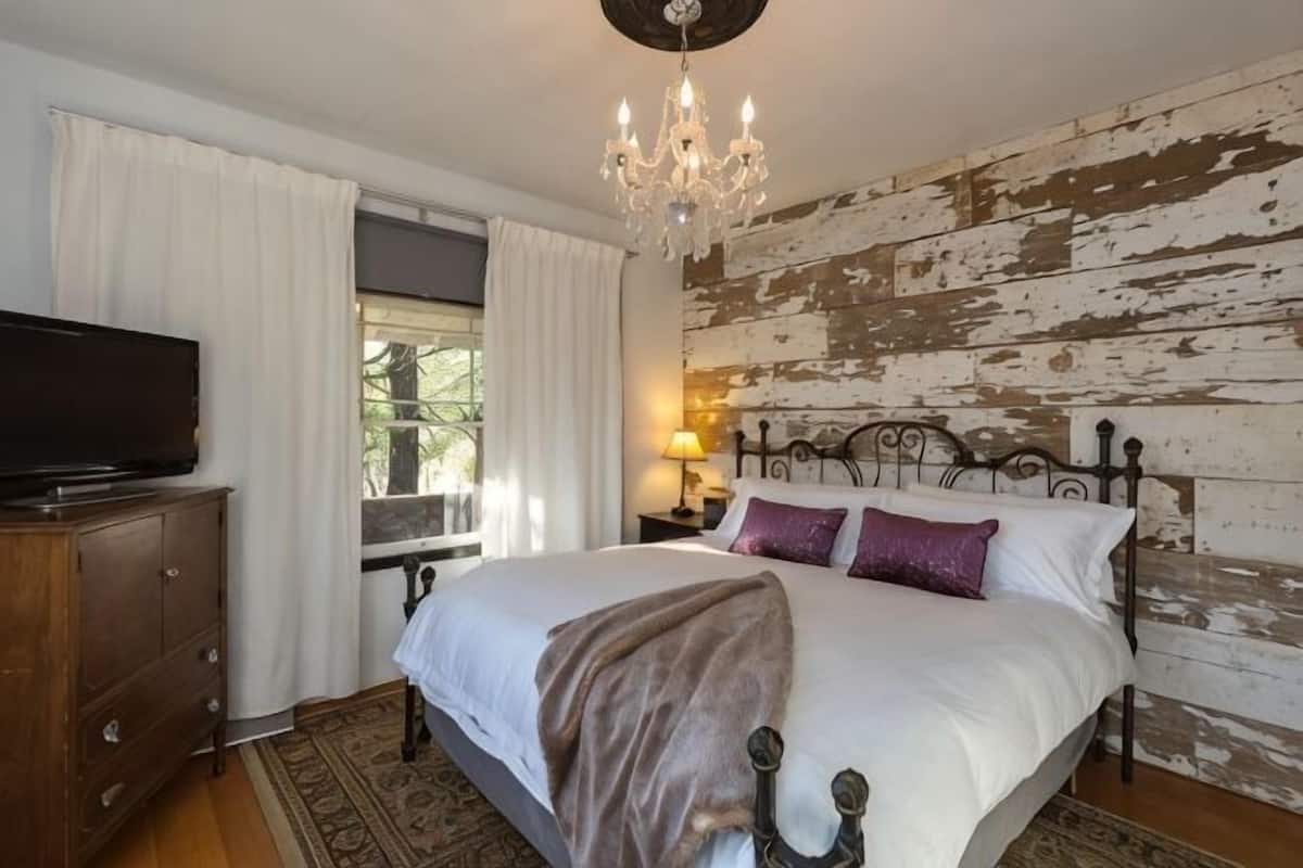 A bedroom with a bed, a television on a dresser, a chandelier, and a rustic wall.