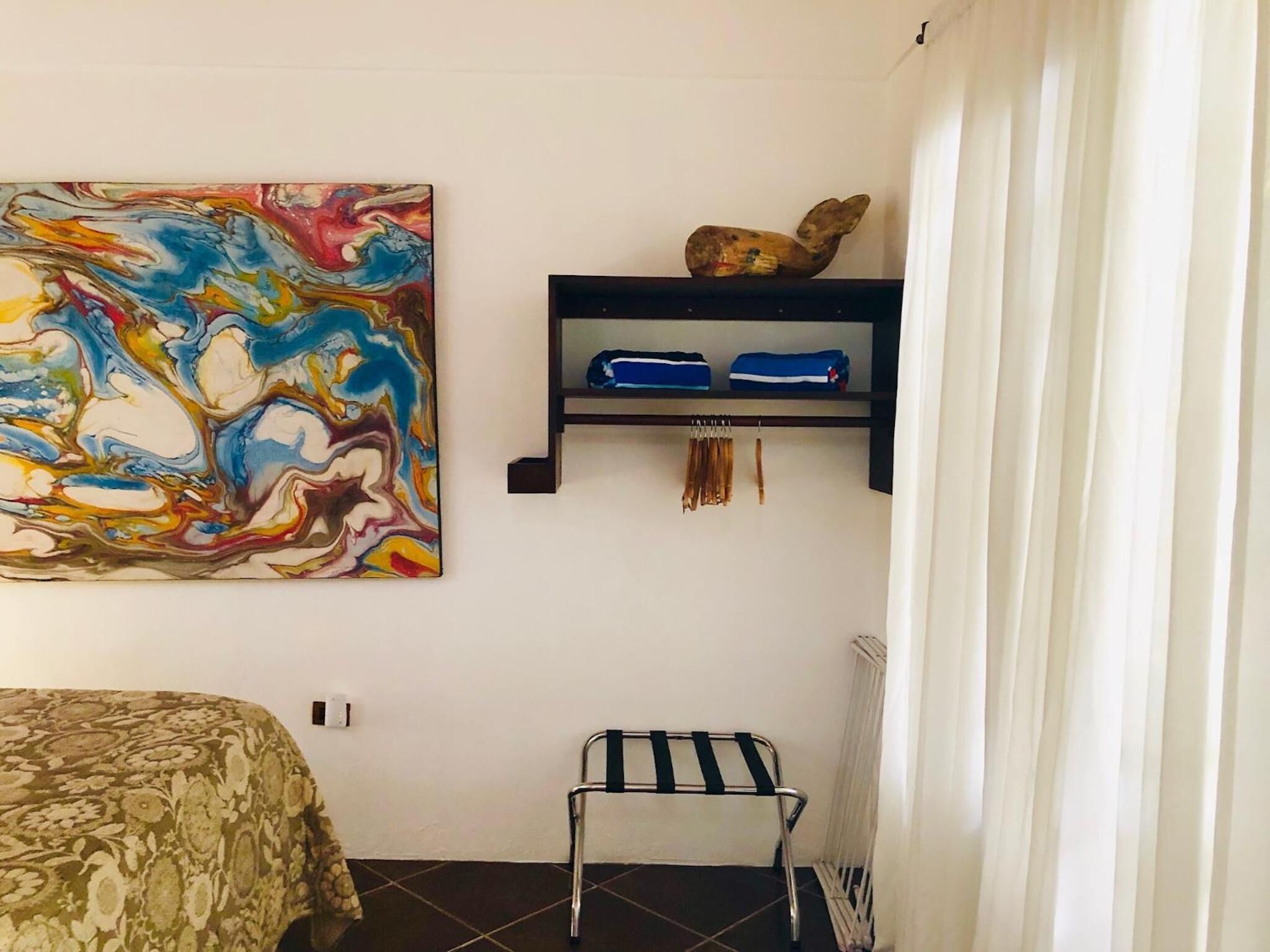 A bedroom with a bed, a chair, a colorful abstract painting, a shelf with folded clothes, and a radiator.