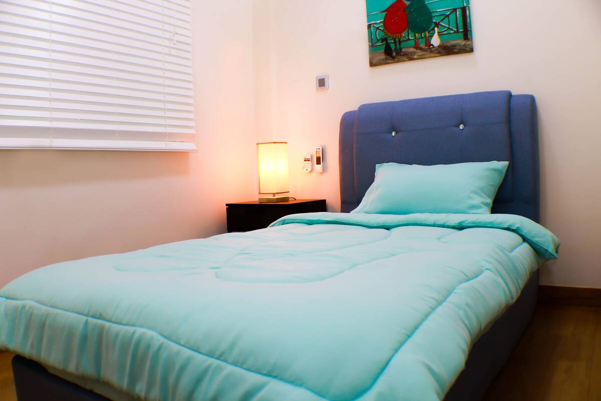A neatly made bed with a light blue comforter and a matching pillow, a bedside lamp, and a small bedside table.