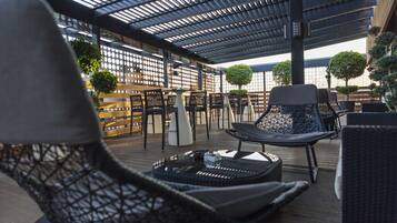 Een terras op het dak met buitenmeubilair, waaronder rieten stoelen en een tafel, onder een metalen dakconstructie.