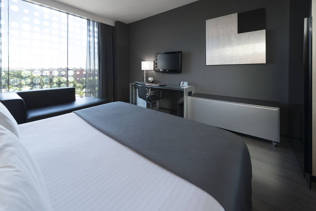 Una habitación de hotel moderna con una cama grande, un escritorio con silla, un televisor de pantalla plana y vistas a la ciudad a través de amplias ventanas.
