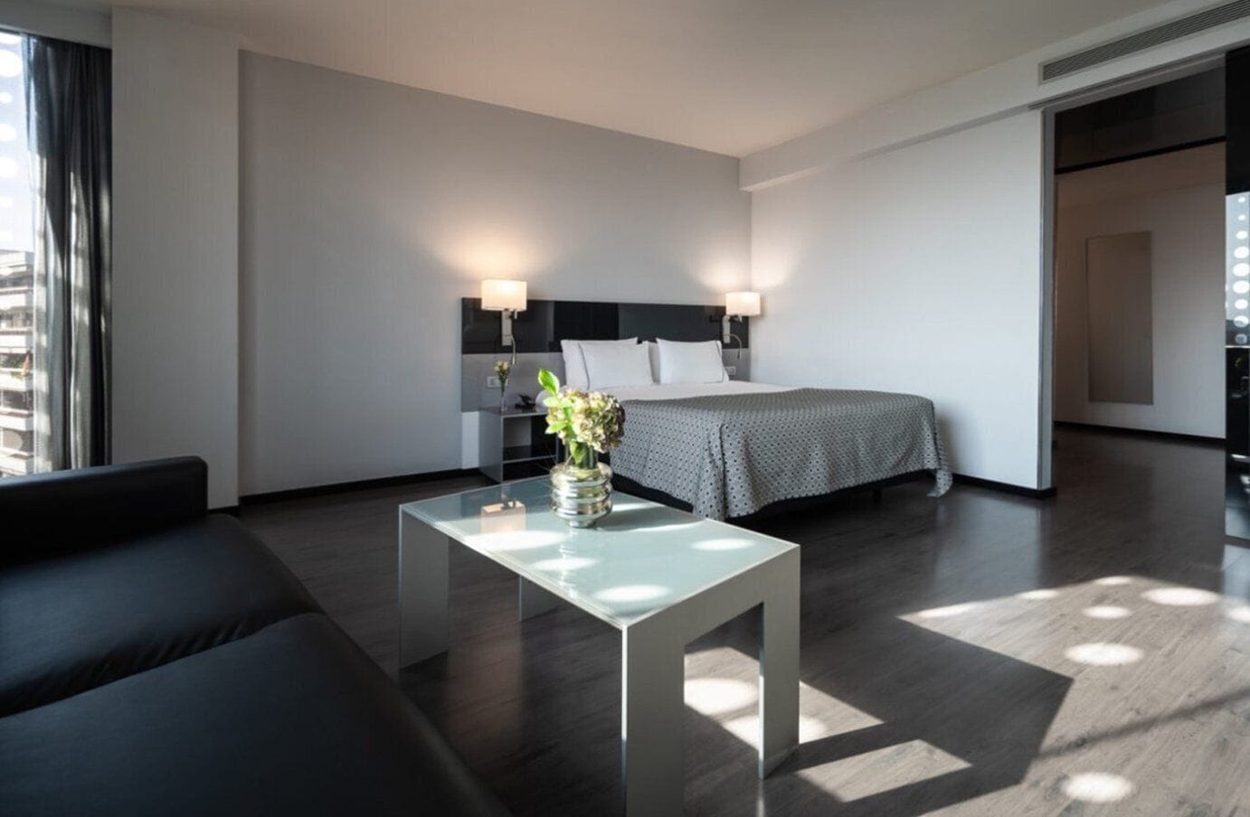 Una habitación de hotel moderna con una cama, mesitas de noche, una mesa de centro de vidrio y un sofá.