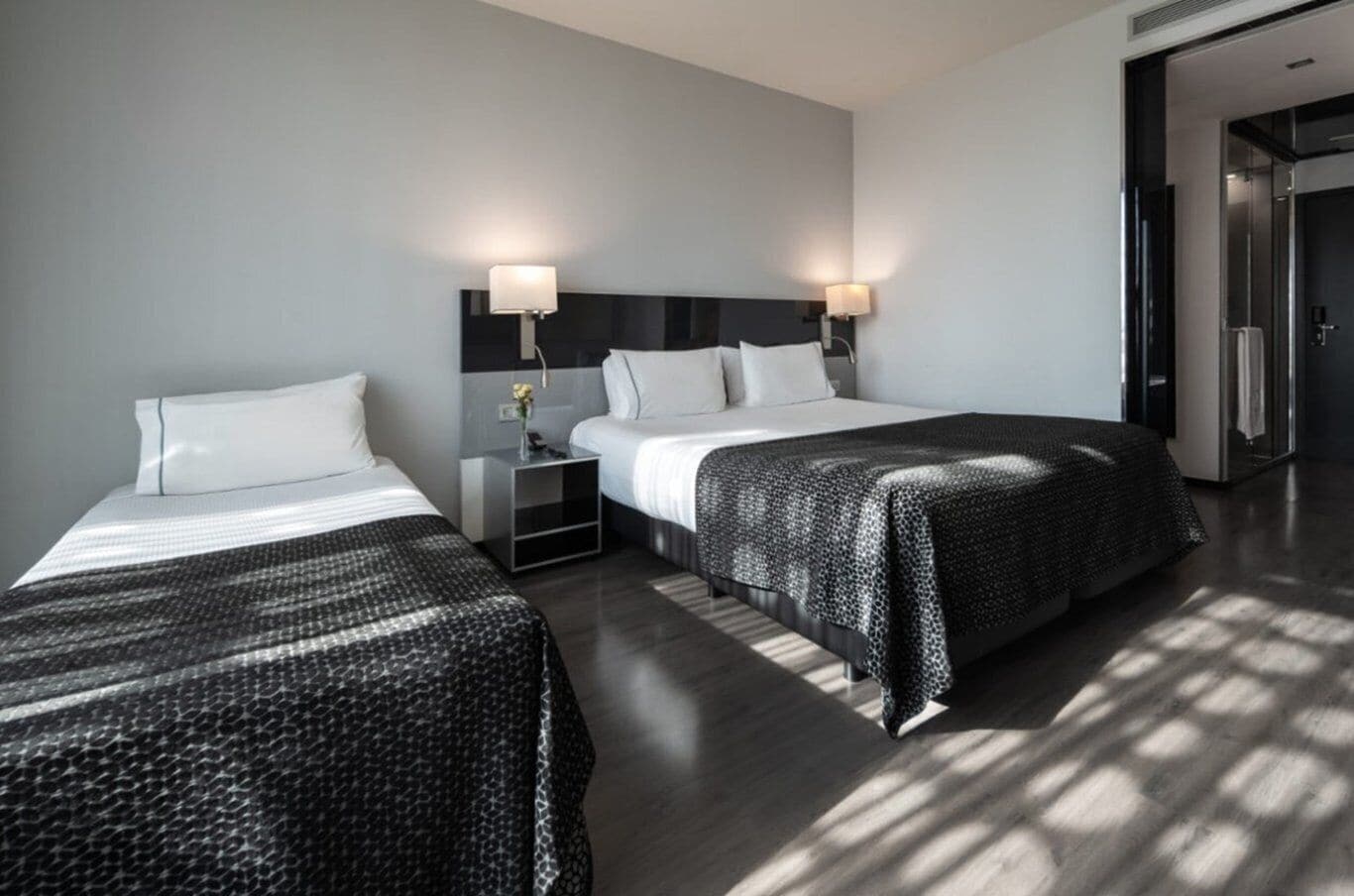A modern hotel room with two beds, a bedside table with a lamp, and a large mirror.