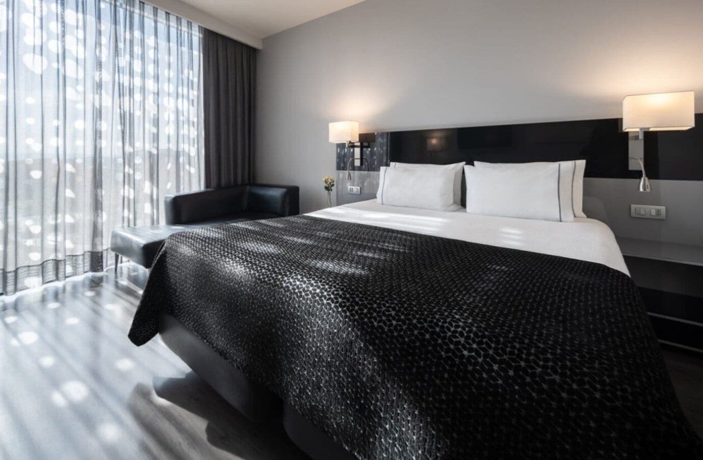 Una habitación de hotel moderna con una cama grande, una silla, una mesita de noche y un ventanal con cortinas.