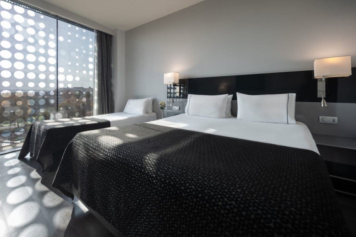 Una camera d'albergo con due letti, un'ampia finestra vista e un comodino con una lampada.