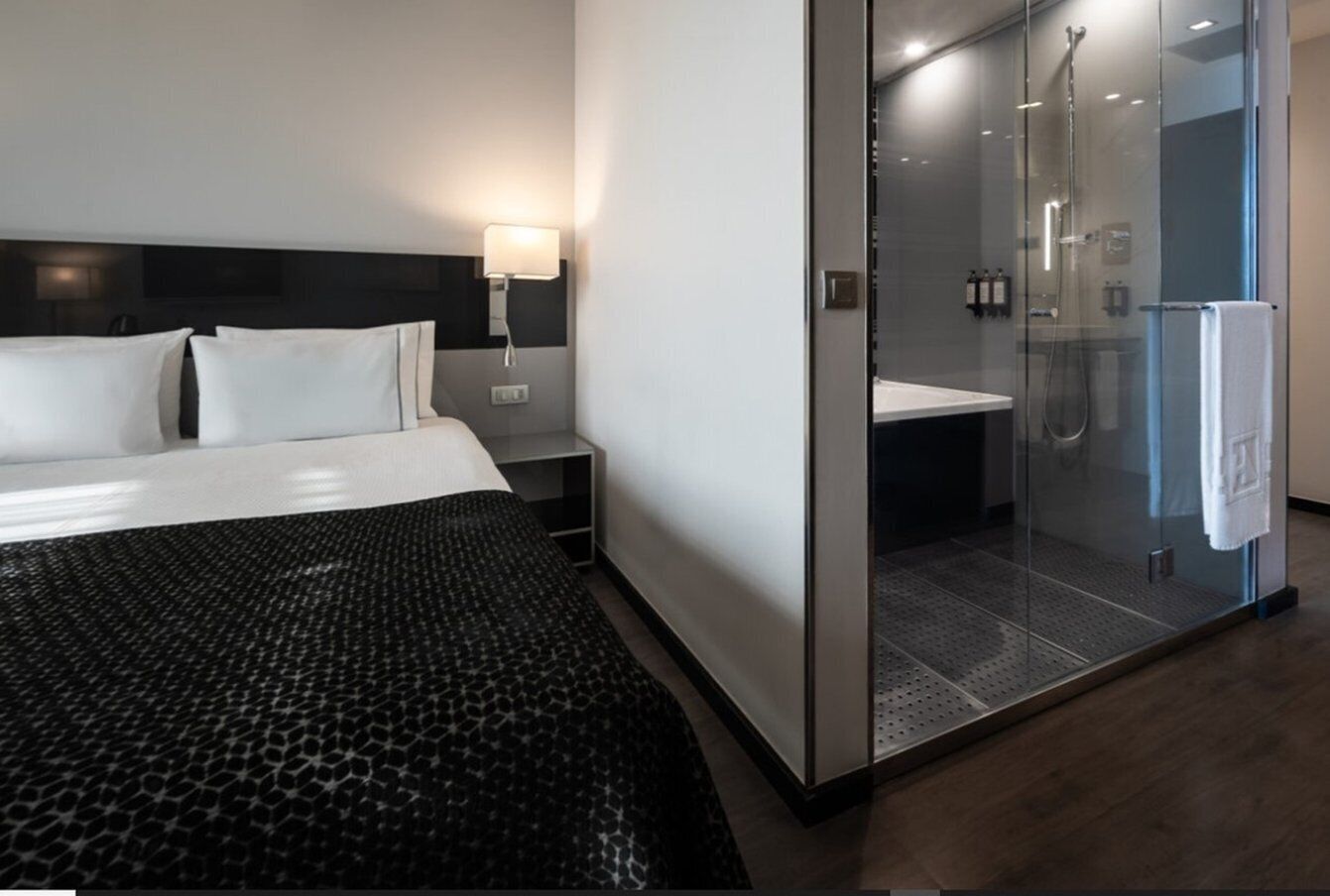 Ein modernes Hotelzimmer mit einem großen Bett, einer Glaskabine für die Dusche und einem Nachttisch mit Lampe.