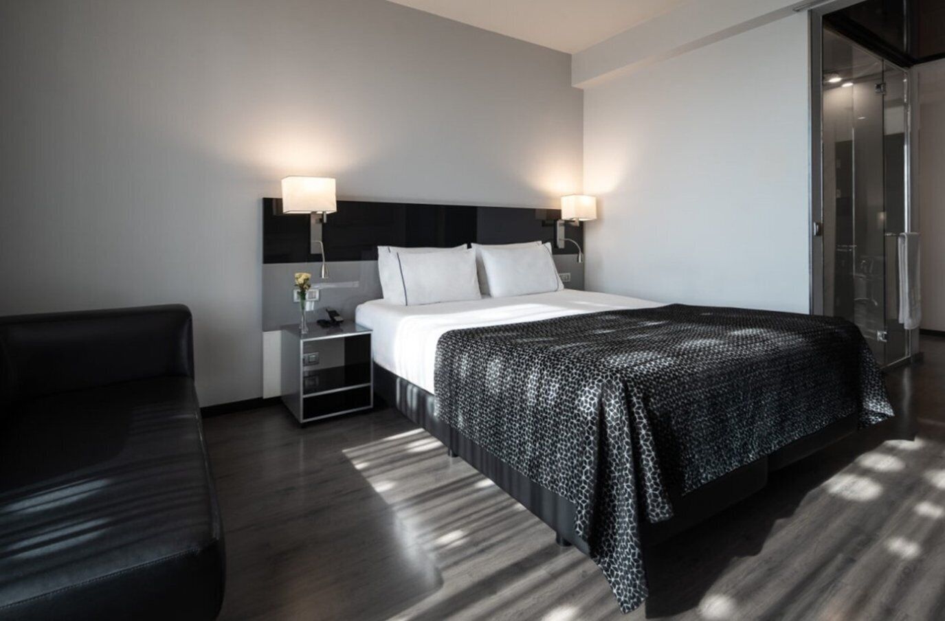 Una habitación de hotel moderna con una cama grande, una mesita de noche, un sofá de cuero negro y un cerramiento de ducha de vidrio.