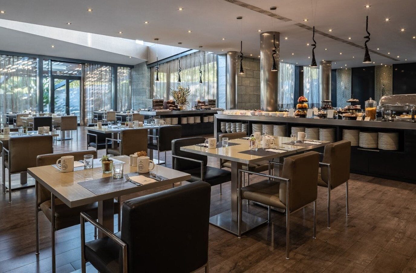 A modern restaurant with multiple dining tables and chairs, a bar area with a buffet, and large windows allowing natural light.