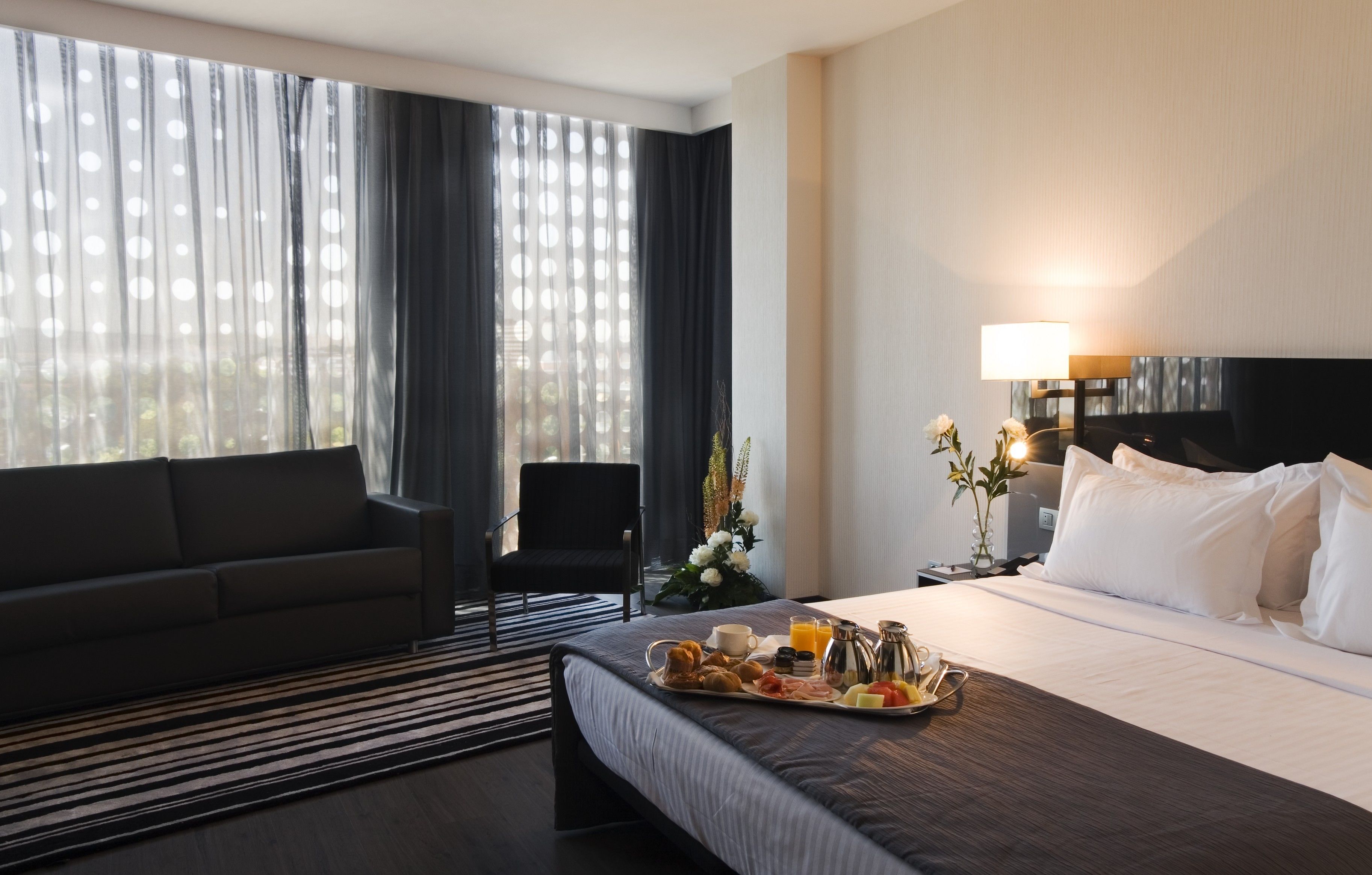 Une chambre d’hôtel comprenant un lit, un canapé, une table à manger avec le petit-déjeuner, une télévision et une fenêtre avec des rideaux.