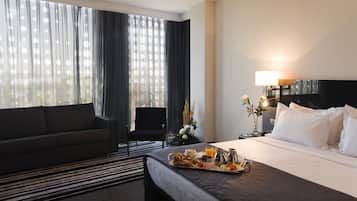 Habitación de hotel con cama, sofá, mesa de comedor con desayuno, televisor y ventana con cortinas.