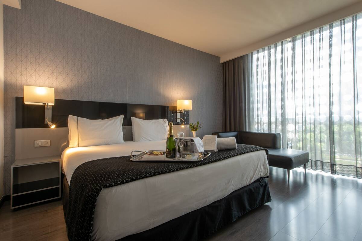 Una habitación de hotel moderna con una cama grande, dos lámparas de noche, un mesita de noche, una zona de estar y un amplio ventanal con cortinas.
