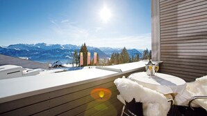 En balkong med utsikt över snöklädda berg, ett bord med en flaska och ett glas, och ett vitt, fluffigt föremål.