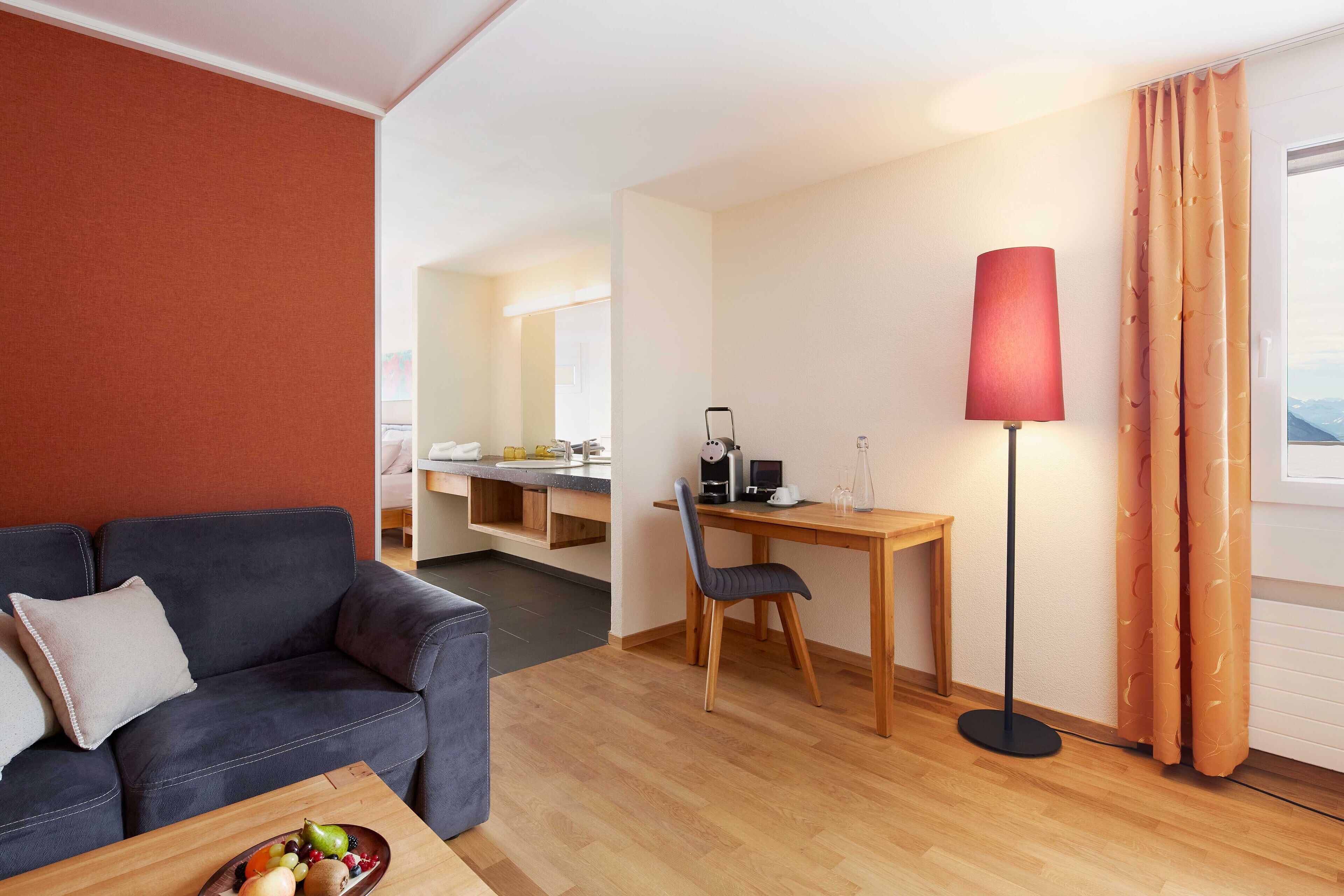Ett modernt hotellrum med en mörkgrå soffa, ett matbord i trä, ett skrivbord med en lampa och en utsikt genom ett fönster med gardiner.