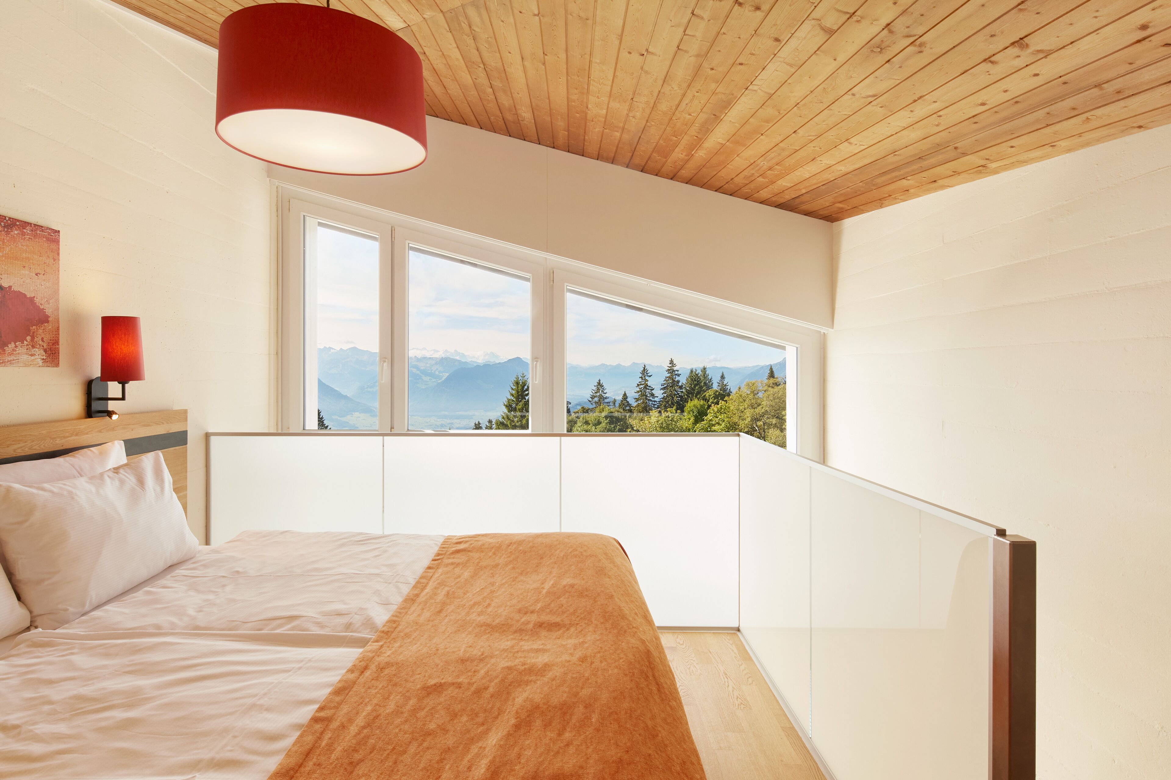 A bedroom with a large bed, a red pendant light, and a view of mountains through a window.
