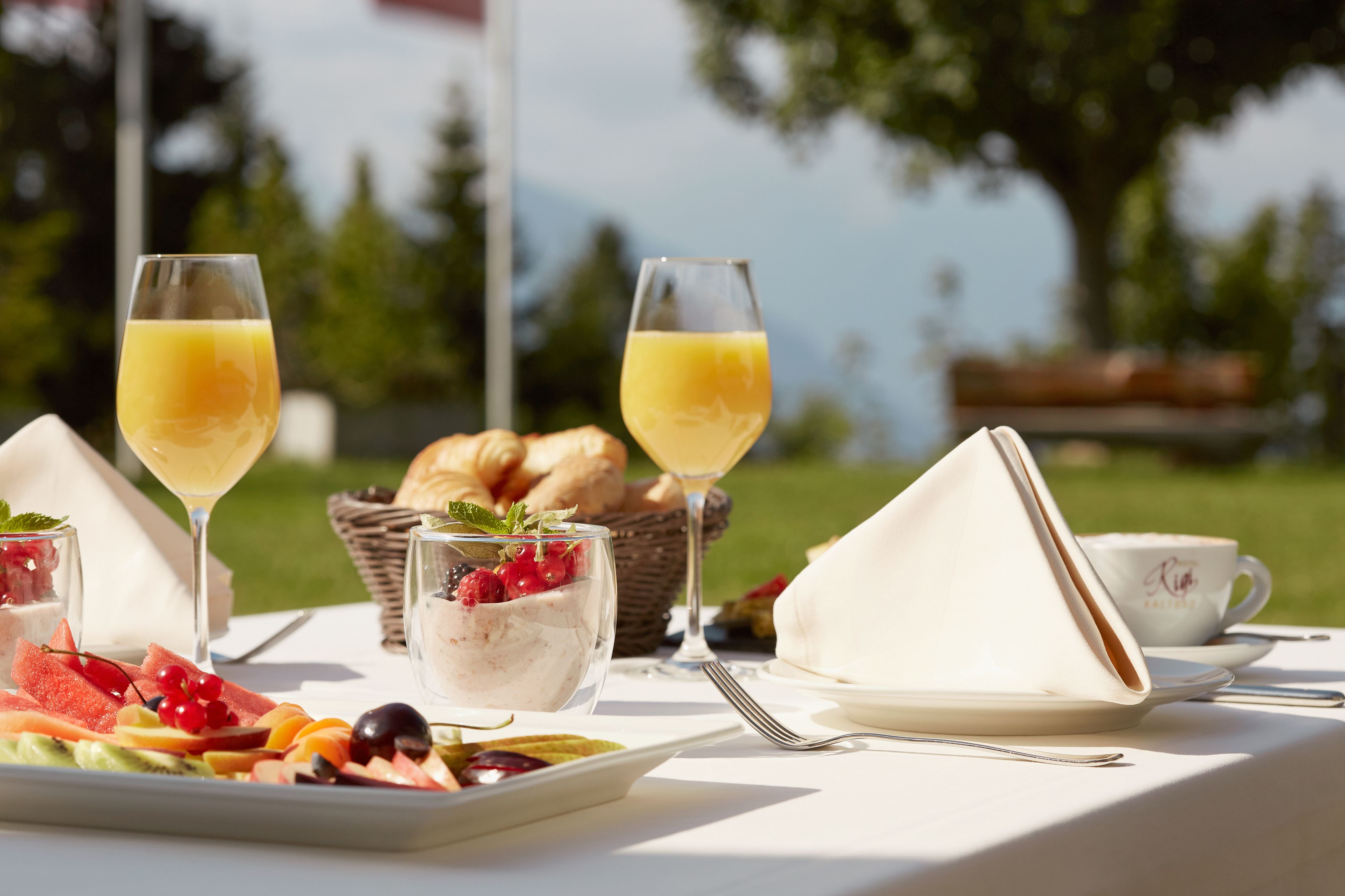 A breakfast setting with a croissant basket, a plate of fruit, and two glasses of orange juice on a table outdoors.