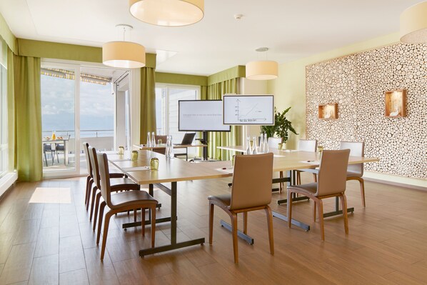 Un salón de conferencias con una gran mesa, sillas, una pantalla de televisión y vistas al mar a través de un ventanal.