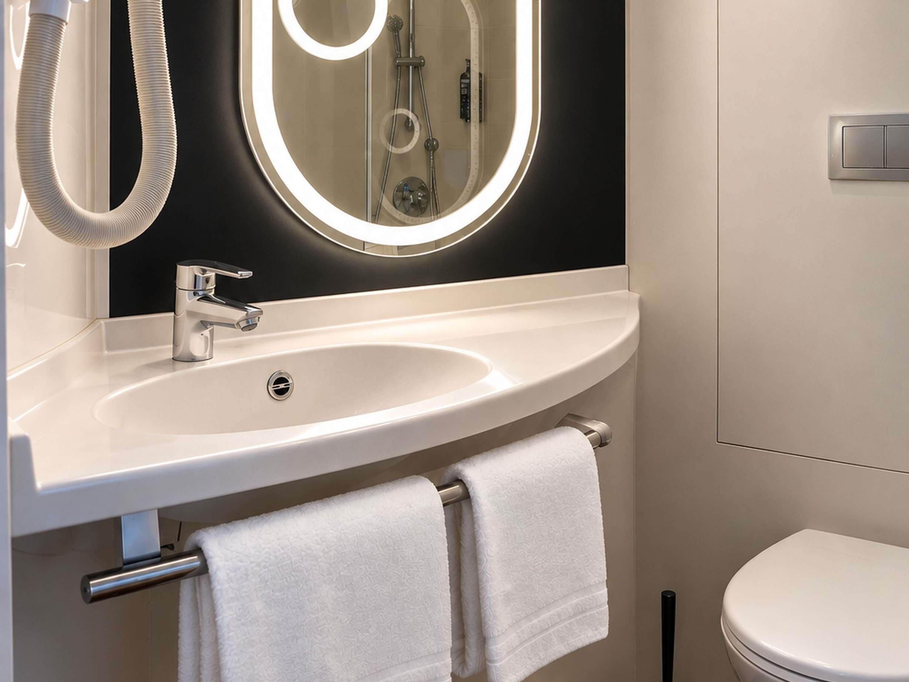 A modern bathroom with a white sink, a mirror with a light halo, a handheld showerhead, and a toilet.