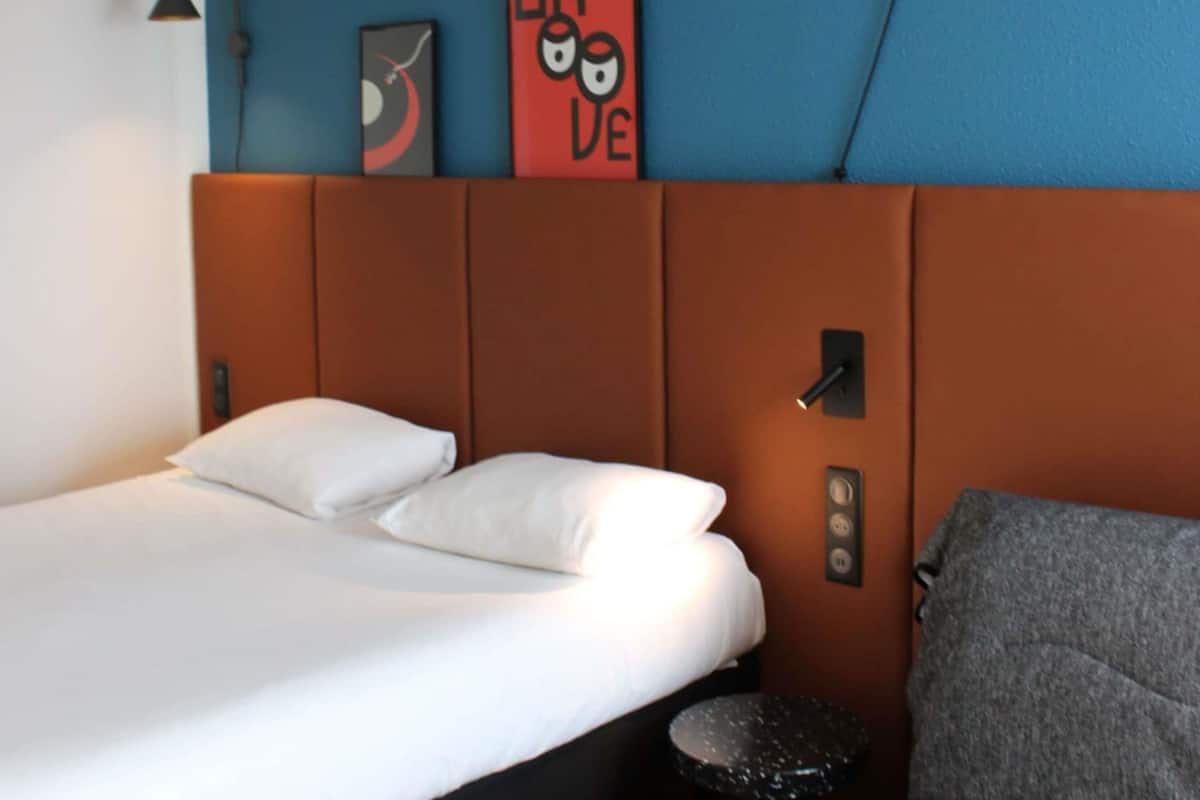 A hotel room with a bed, a small round table, a chair, a wall-mounted light, and a red sign with a cartoon character.