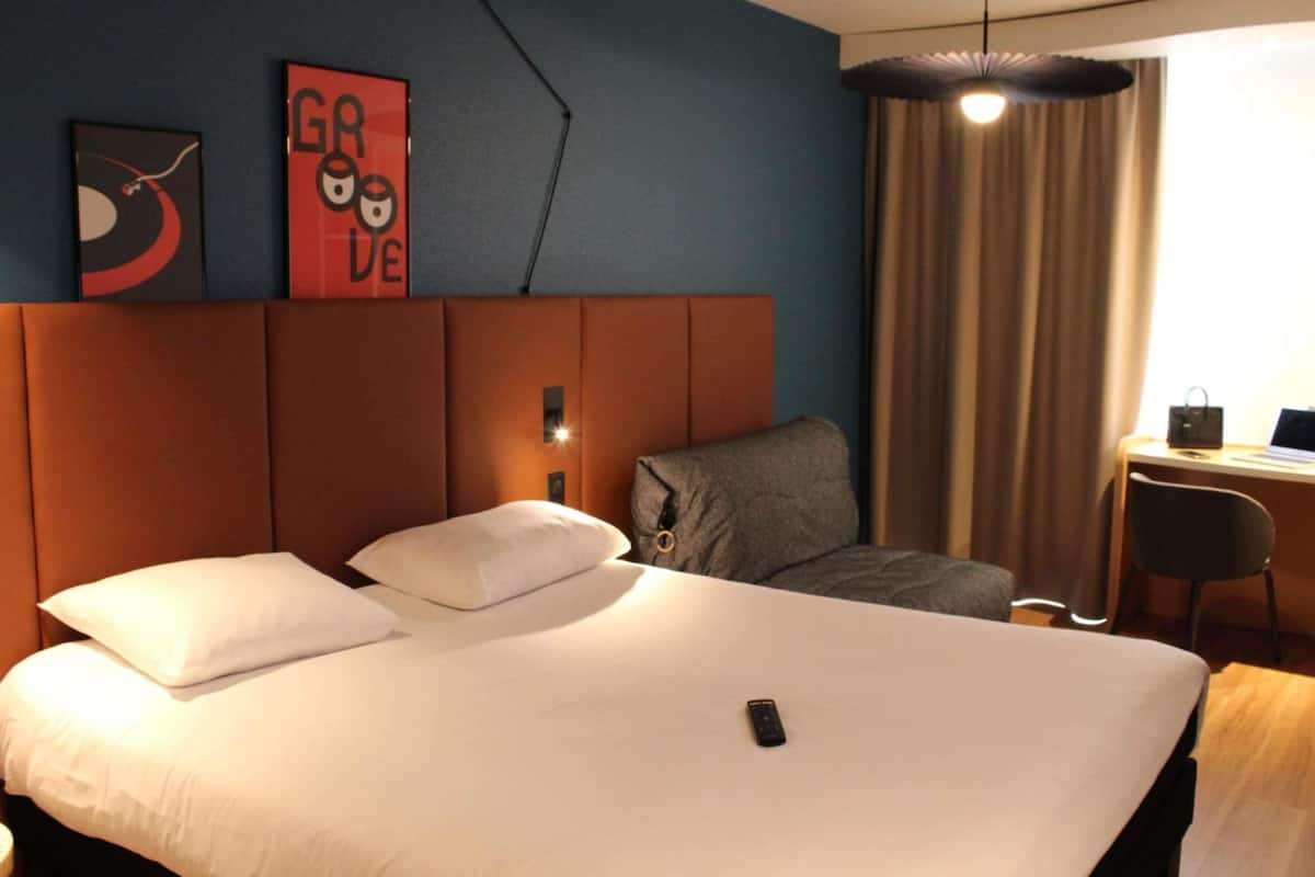 A hotel room with a large bed, a desk, a chair, a lamp, and a wall with a red sign.