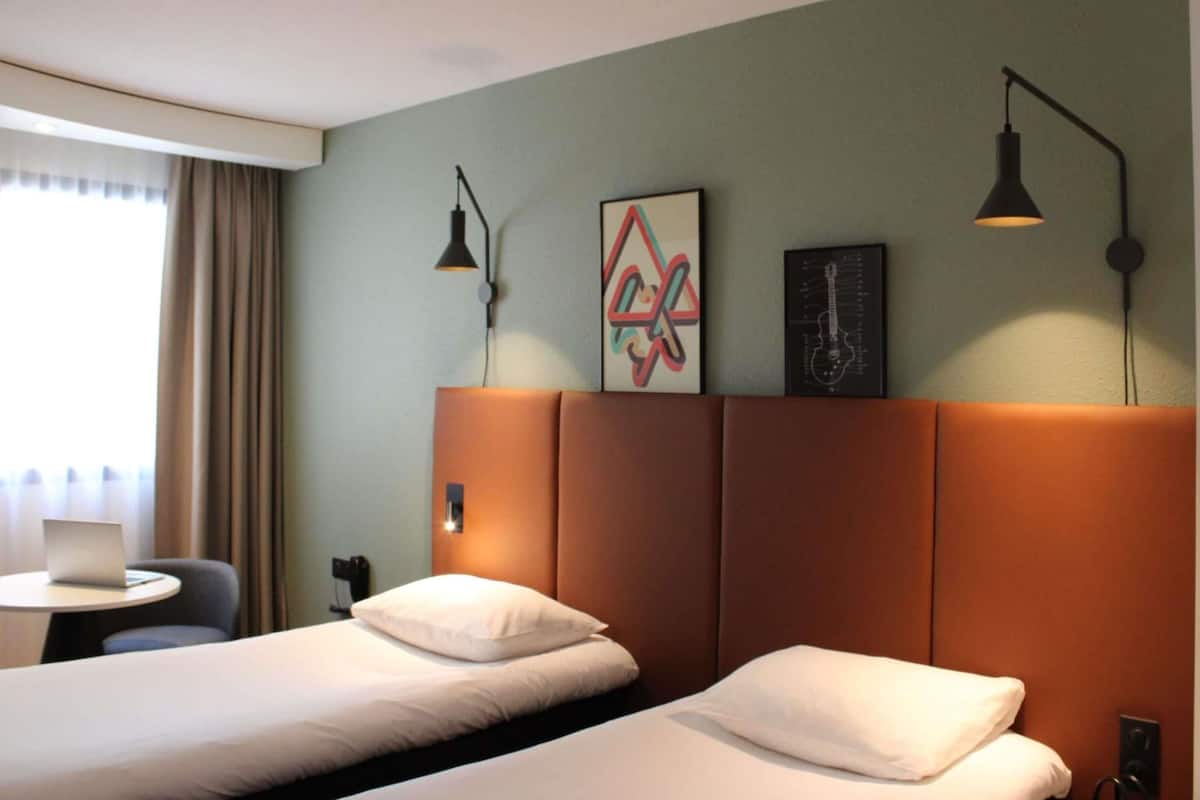 A hotel room with two beds, a desk, a chair, and artwork on the wall.