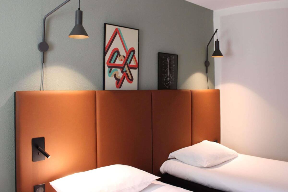 A hotel room with a brown headboard, two beds, a wall-mounted artwork, and two wall-mounted lamps.
