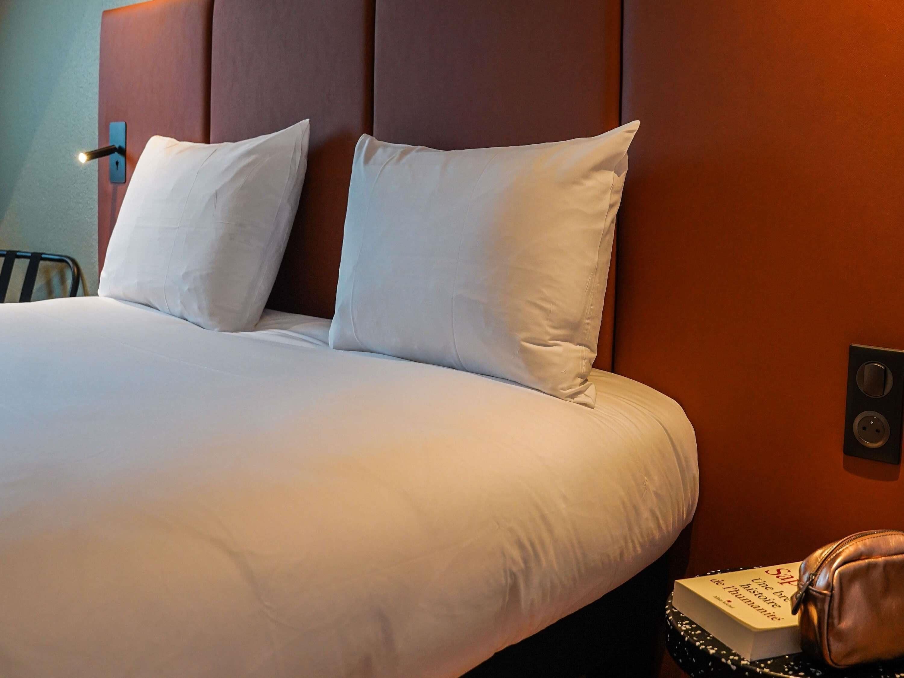A neatly made bed with white linens and pillows, a brown headboard, and a small table with a book and a leather bag.
