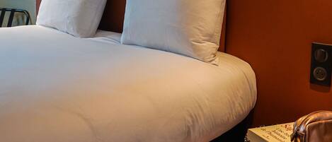 Una cama bien tendida con sábanas y almohadas blancas, un cabecero marrón y una mesita de noche con un libro y un bolso de cuero.