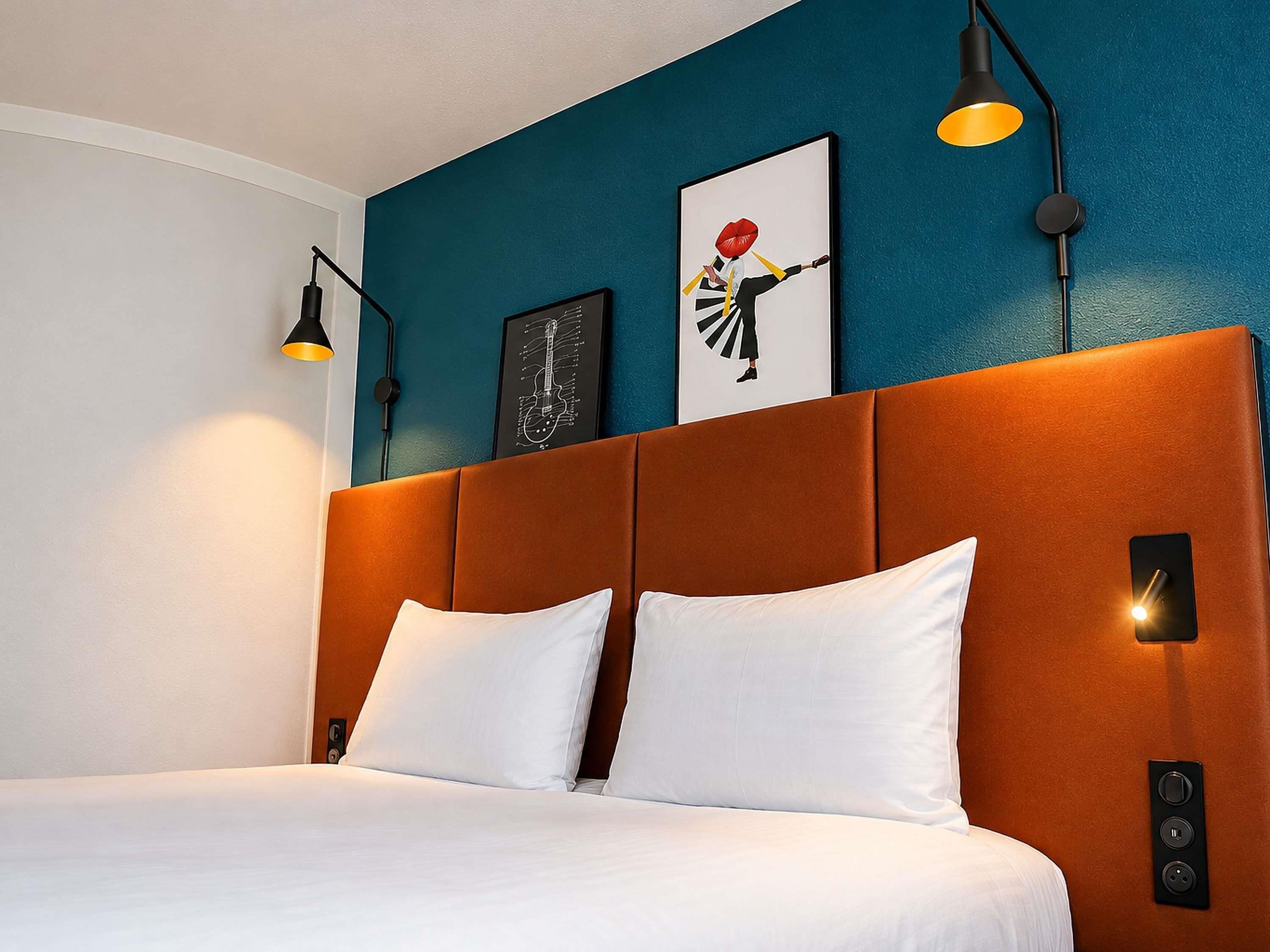 Une chambre d’hôtel avec un lit, une tête de lit, des œuvres d’art murales et un luminaire fixé au mur.