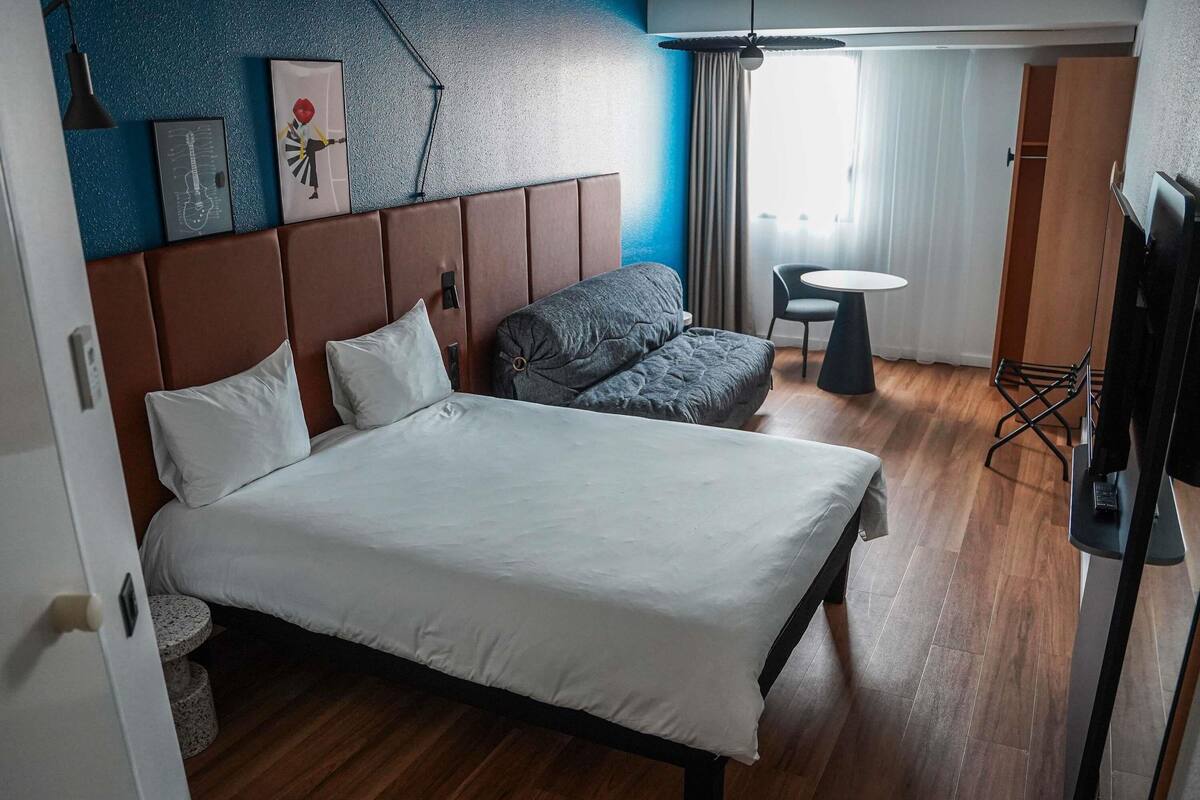 A hotel room with a bed, a sofa, a small table, a TV, and a window with curtains.