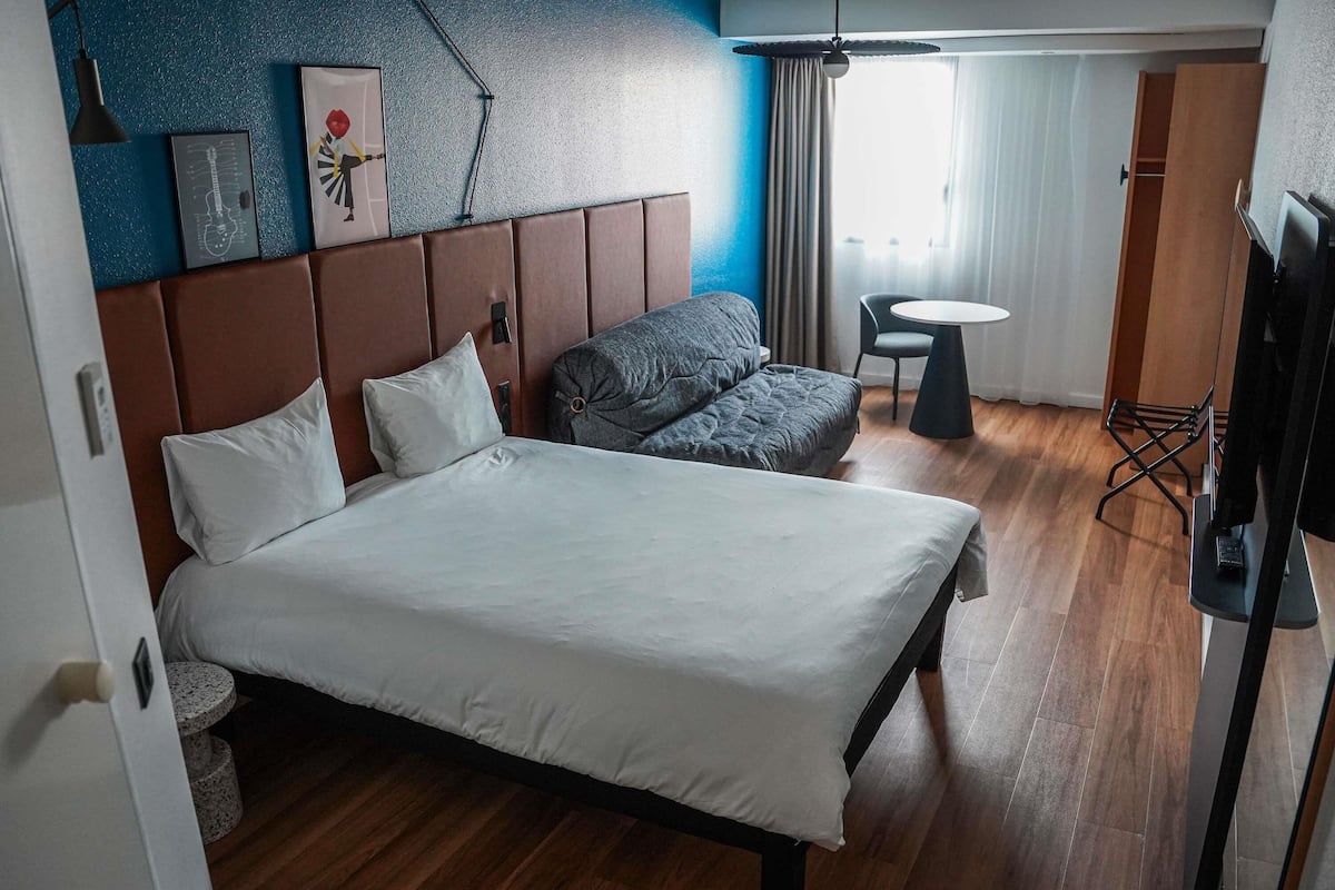 Habitación de hotel con cama, sofá, una mesita, televisor y ventana con cortinas.