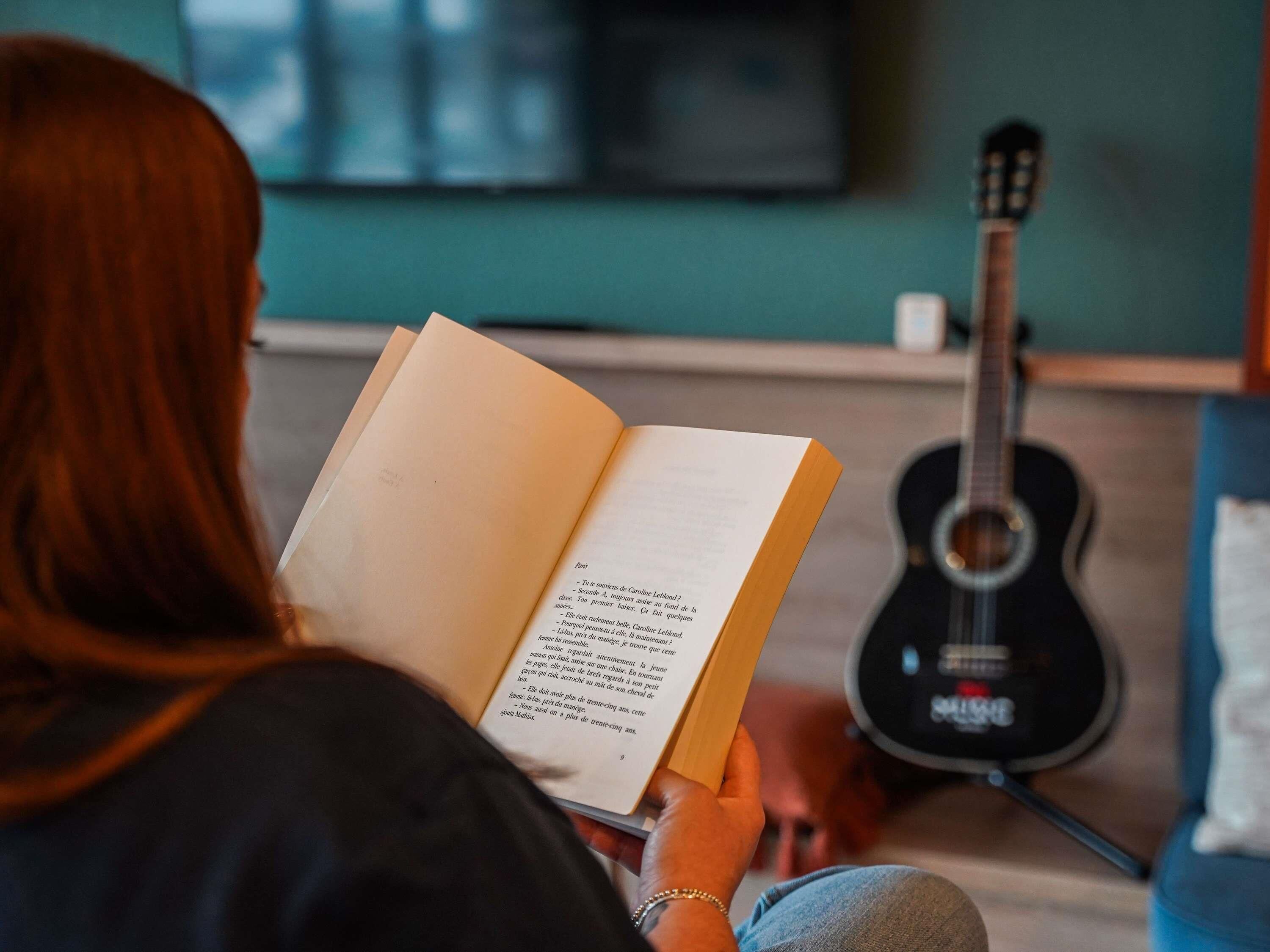 A person reading a book in a room with a guitar and a television in the background.