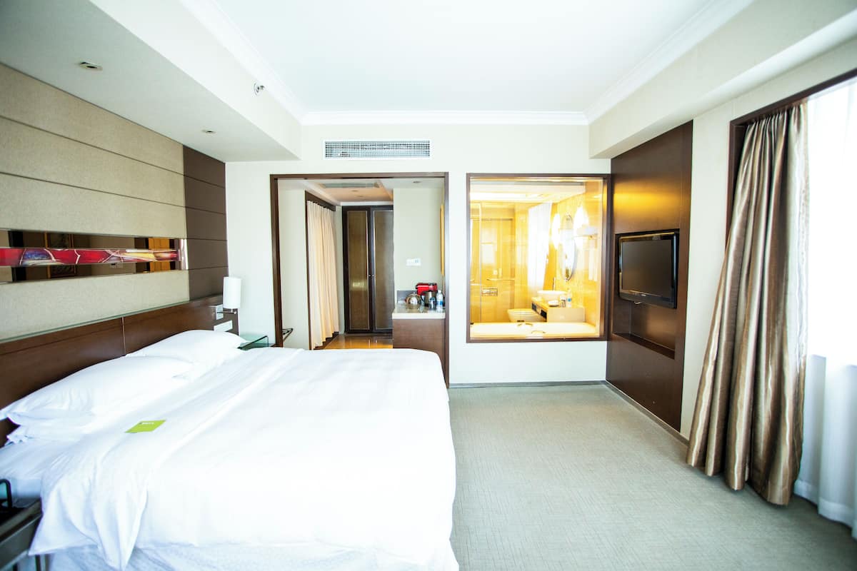 Quarto de hotel com uma cama grande, televisão de tela plana, banheiro com espelho e pia, e vista para o saguão do hotel.