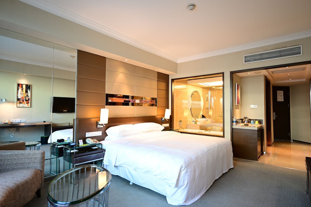 Quarto de hotel com uma cama, uma escrivaninha, uma cadeira, um espelho e um banheiro visível através de uma porta aberta.
