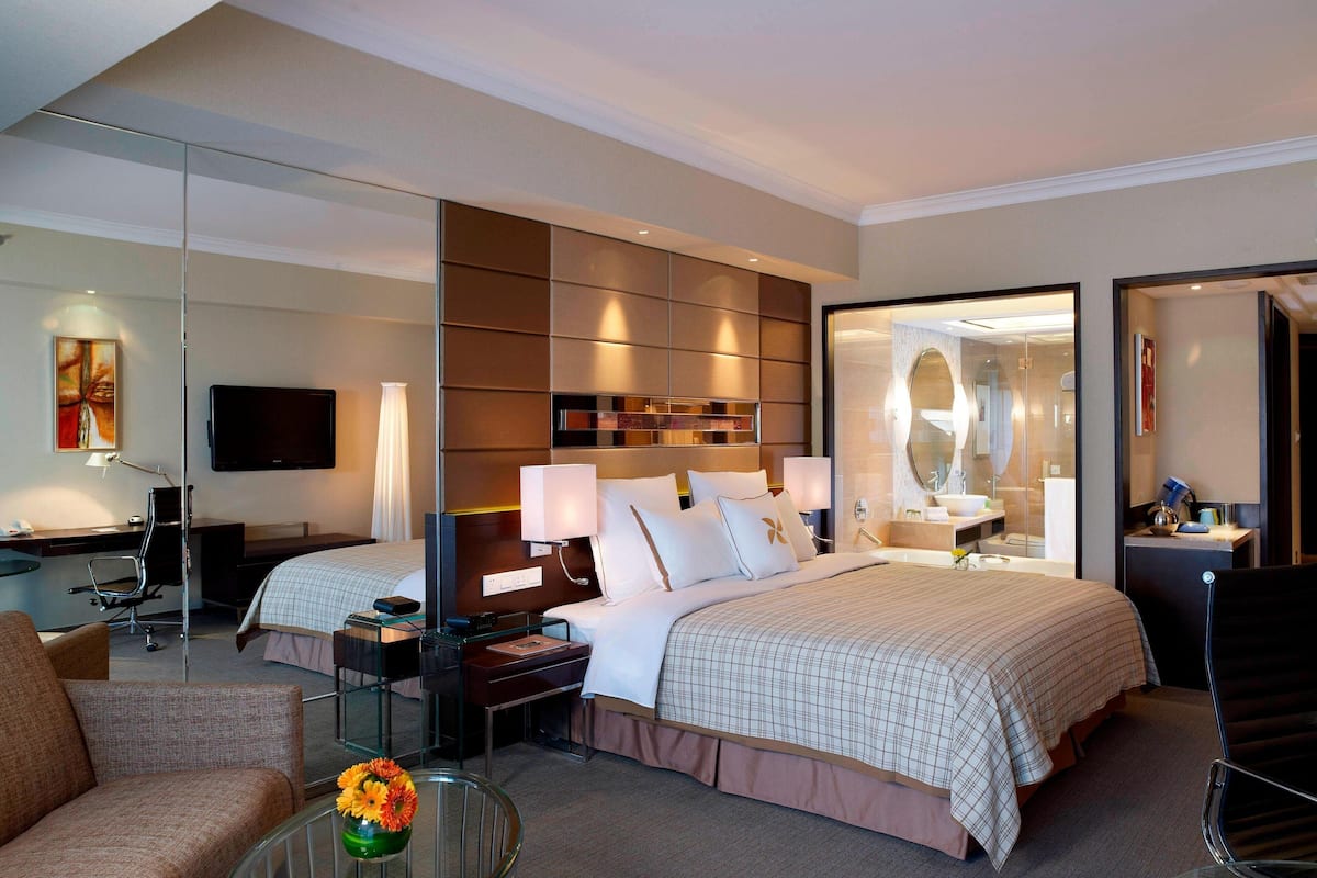 Quarto de hotel moderno com uma cama grande, uma escrivaninha, uma cadeira, uma televisão e um banheiro visível por uma porta aberta.