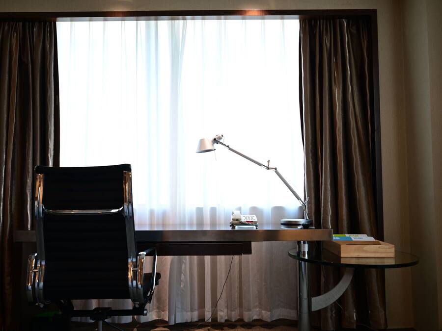 Habitación de hotel con escritorio, silla, lámpara y vistas a través de cortinas translúcidas.