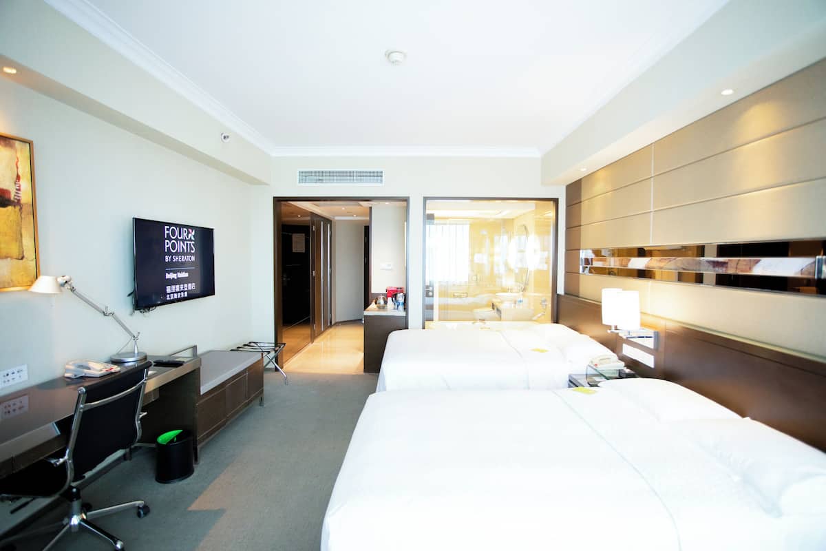 Quarto de hotel com duas camas, uma escrivaninha, televisão e banheiro visível por uma porta de vidro.