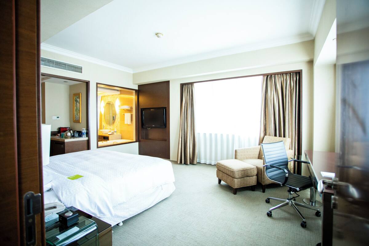 Quarto de hotel com uma cama grande, uma escrivaninha com computador, uma cadeira, uma televisão e uma janela com cortinas.