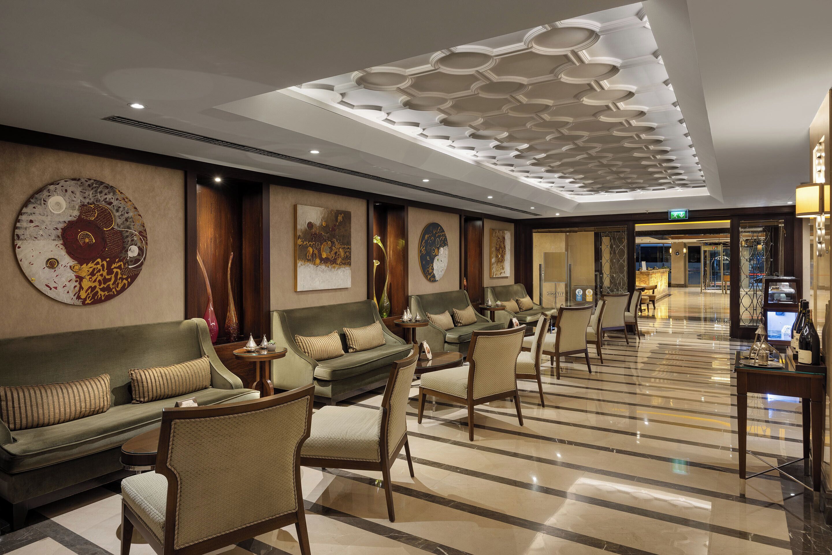 Un hall d’hôtel au design moderne, comprenant un espace salon avec des canapés et des fauteuils, un sol à motifs et des panneaux décoratifs au plafond.