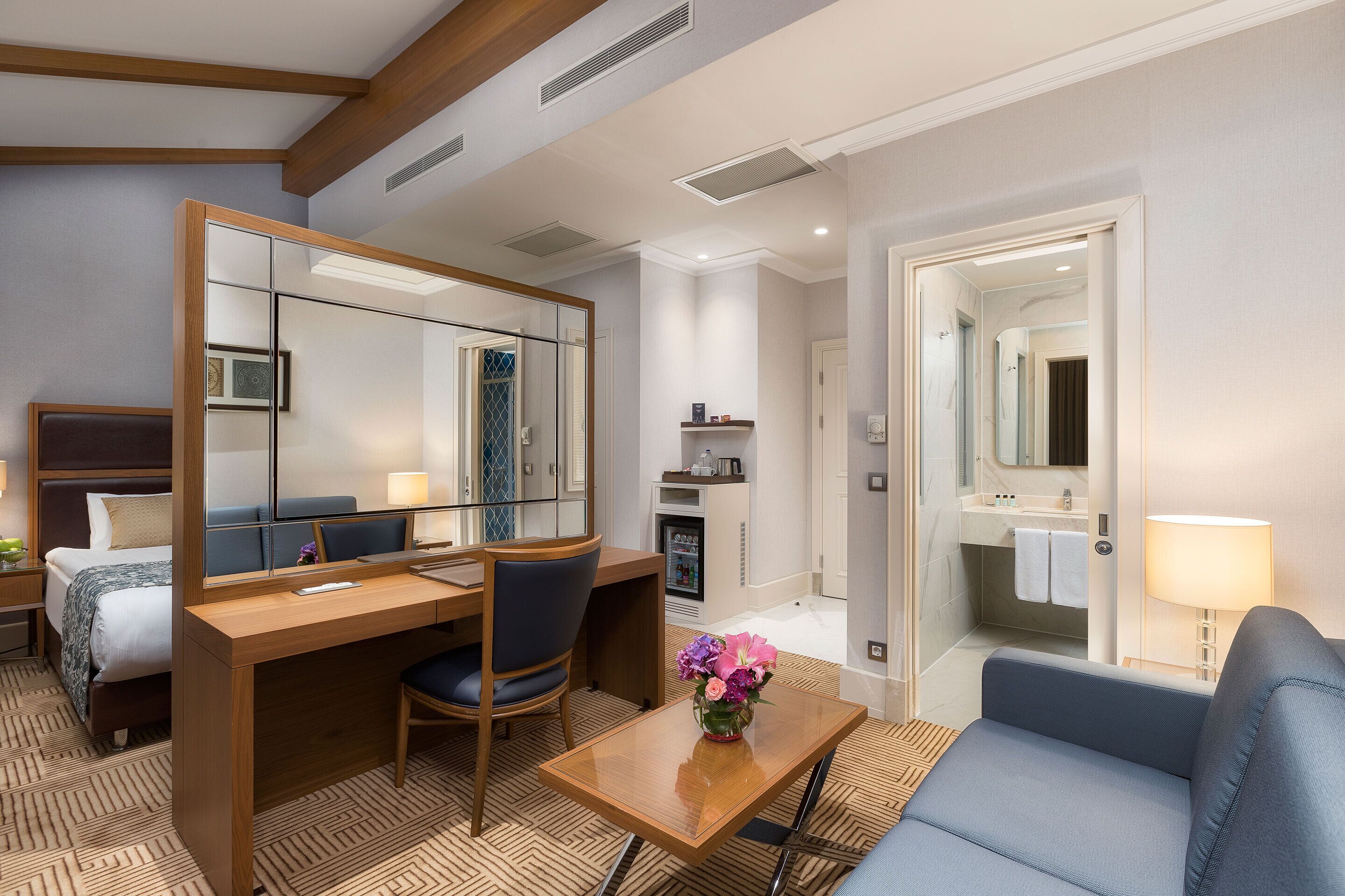 A hotel room with a bed, a desk, a chair, a sofa, a small table with a vase, a mirror, a bathroom with a sink and a towel rack.