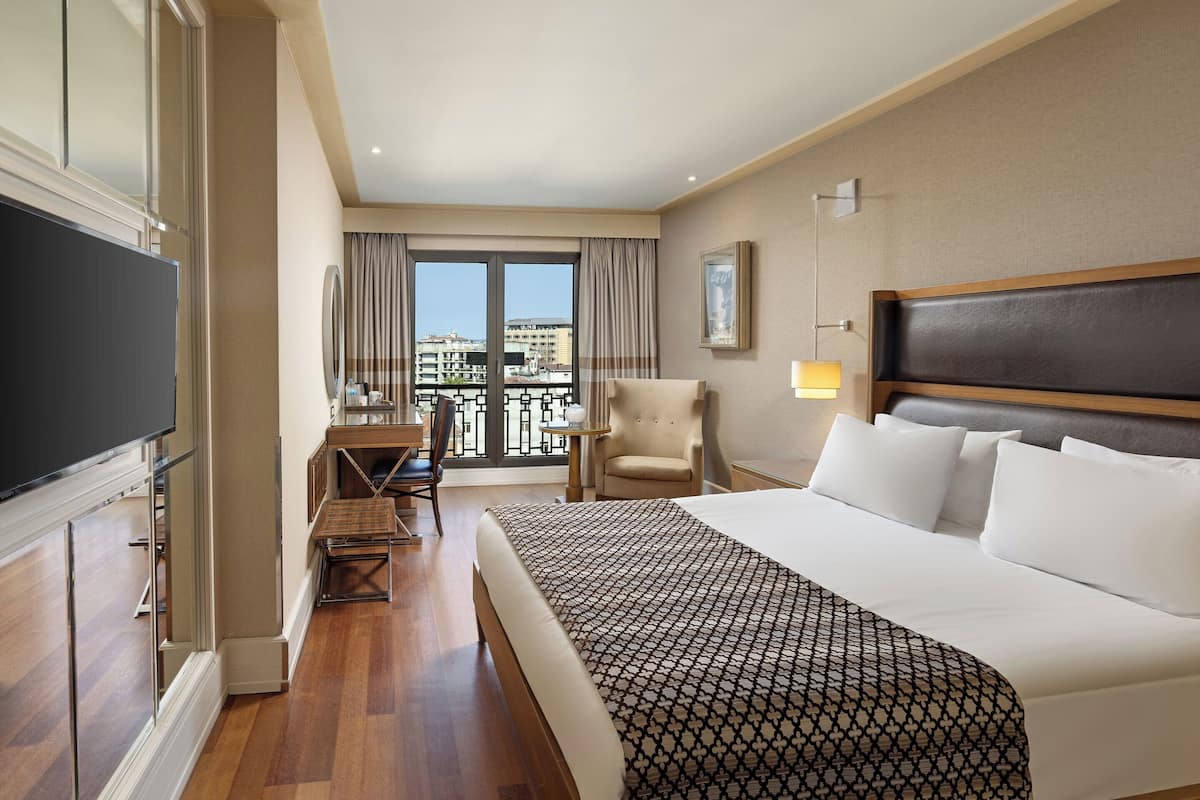 Habitación de hotel con una cama grande, un escritorio, una silla, televisión y balcón con vistas a edificios.