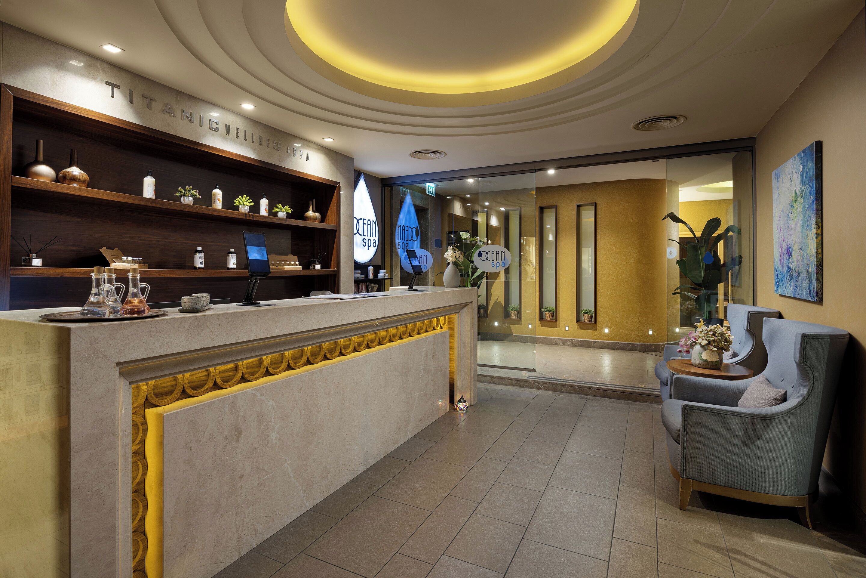 Un'area di accoglienza moderna per la spa, con reception, scaffali per articoli da toilette e un angolo relax con una poltrona e un tavolino.