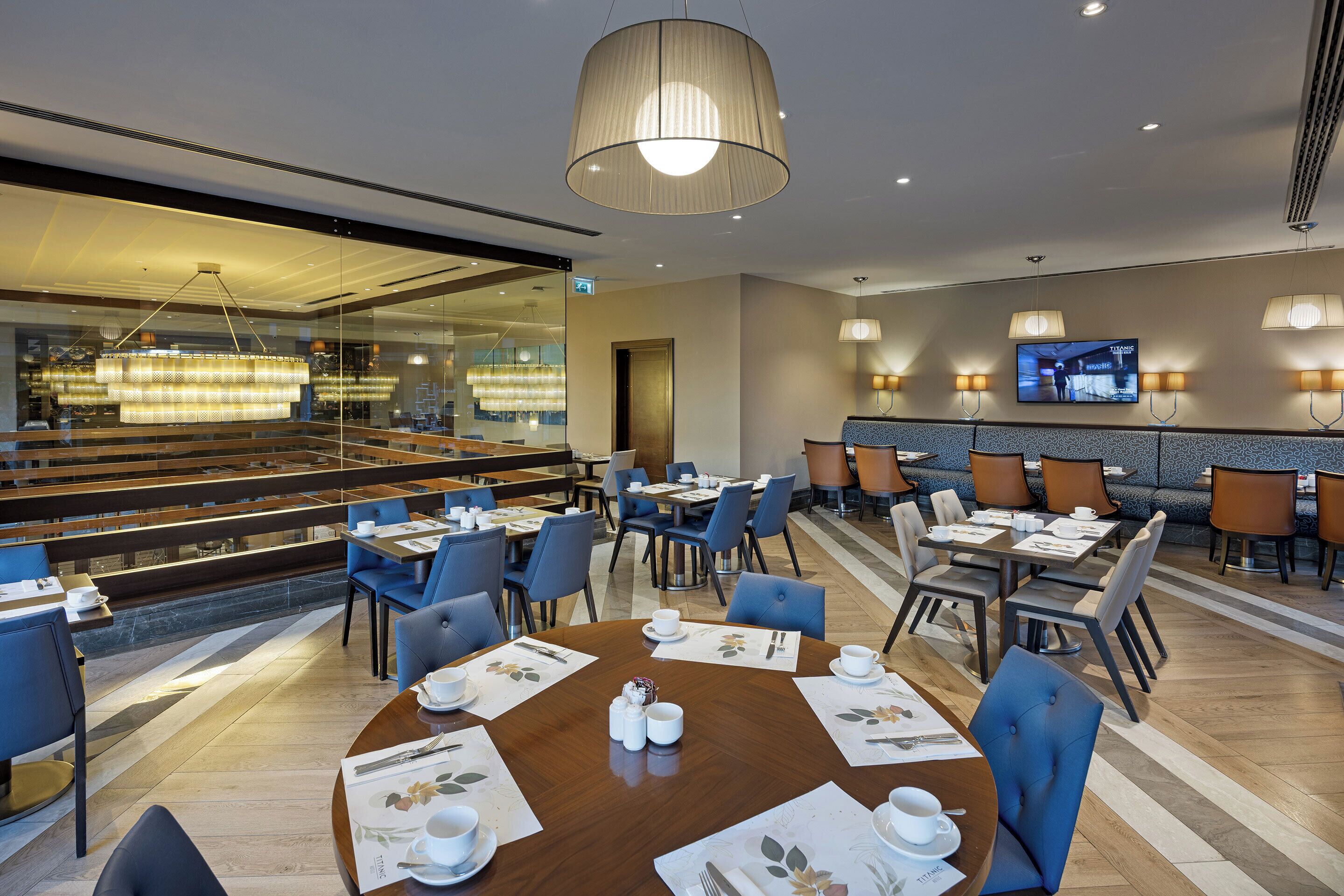 A hotel dining area with round tables, blue chairs, and a large window overlooking a dining area.