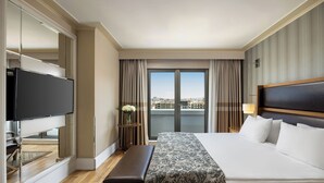 A hotel room with a large bed, a television, a bench, and a view of the city through large windows. - Titanic City Taksim (Istanbul)