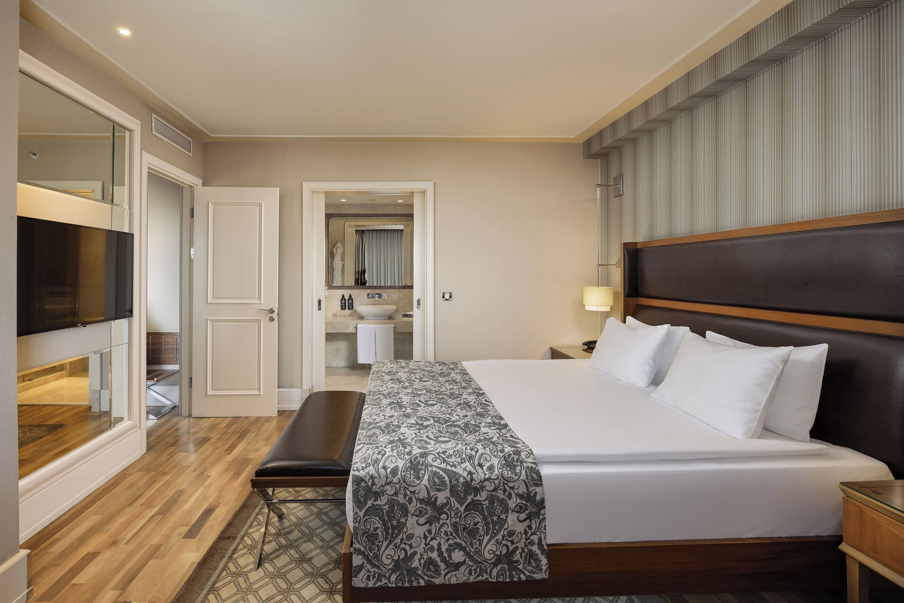Quarto de hotel com uma cama grande, um banco, uma TV e um banheiro visível através de uma porta aberta.