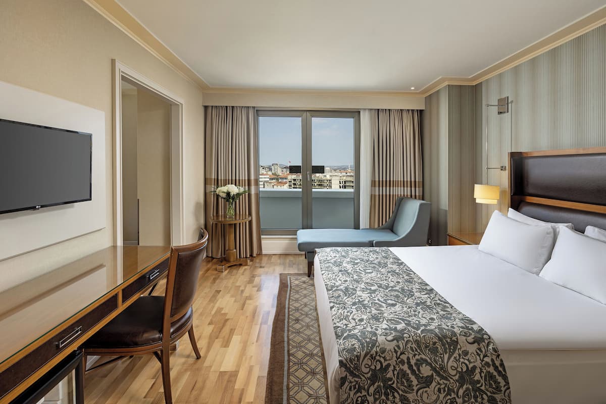 Habitación de hotel con cama, escritorio, silla, televisor y balcón con cortinas, con vista a la ciudad.