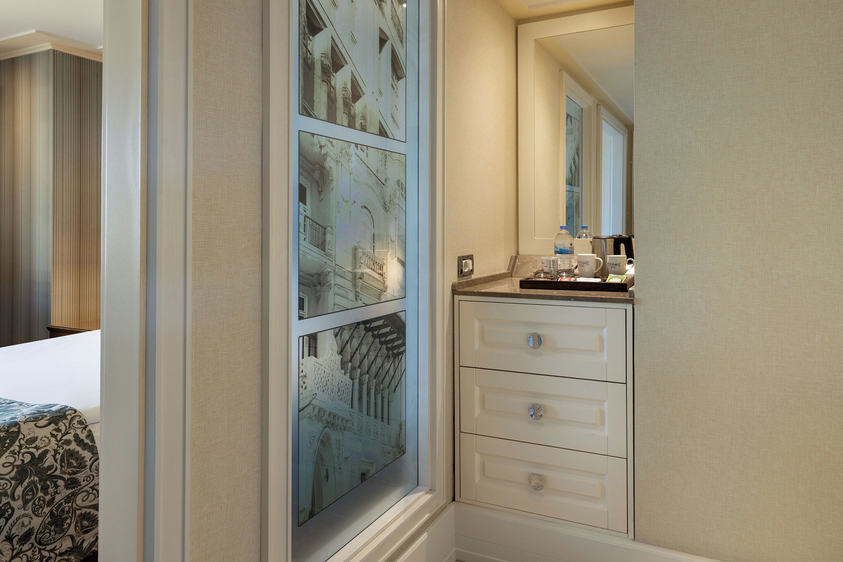 A hotel room with a large window, a bed with a patterned bedspread, a white dresser with drawers, and a mirror above the dresser.