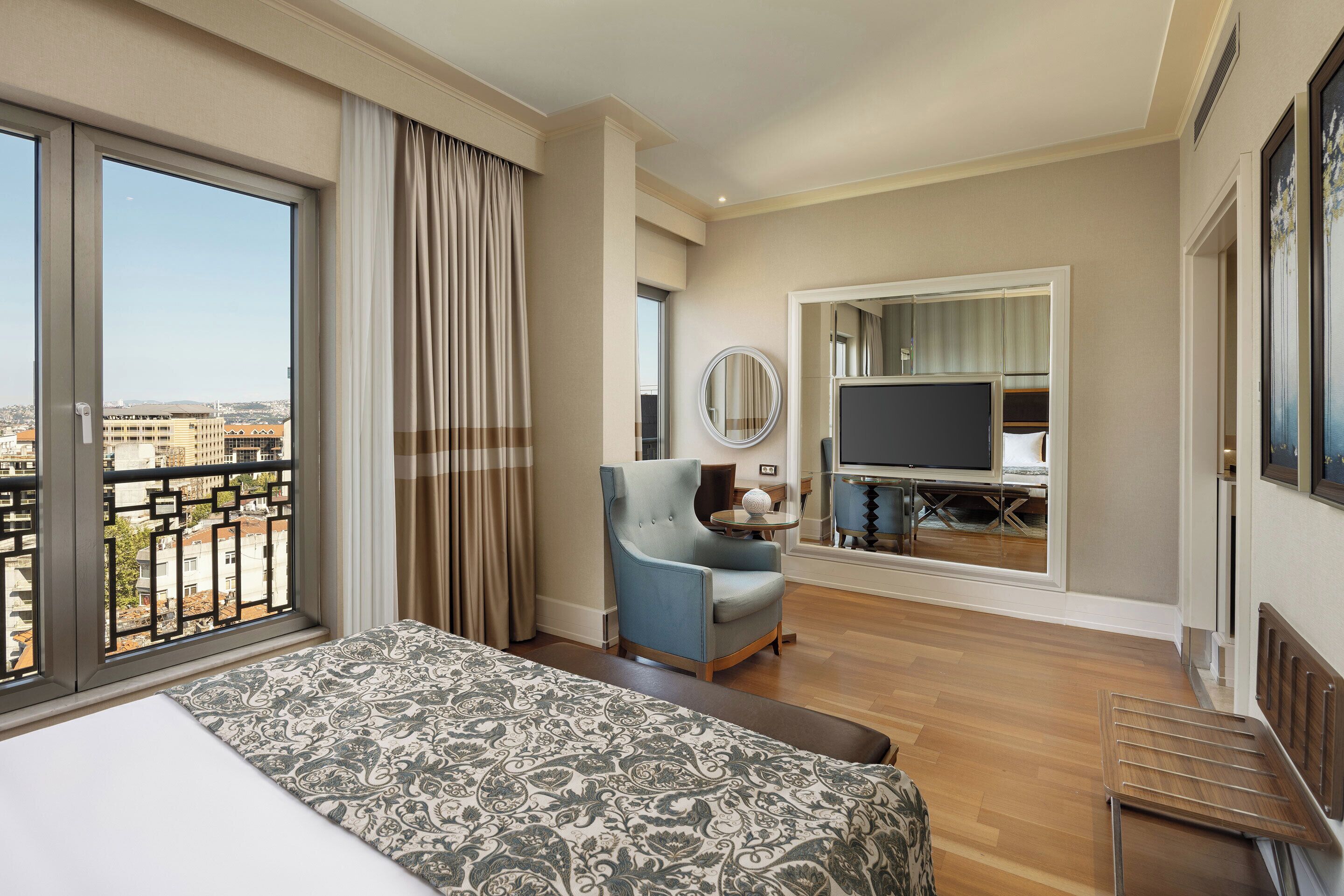 Ein Hotelzimmer mit einem großen Bett, einer Sitzecke mit Sessel und Fernseher sowie einem Balkon mit Blick auf die Stadt.