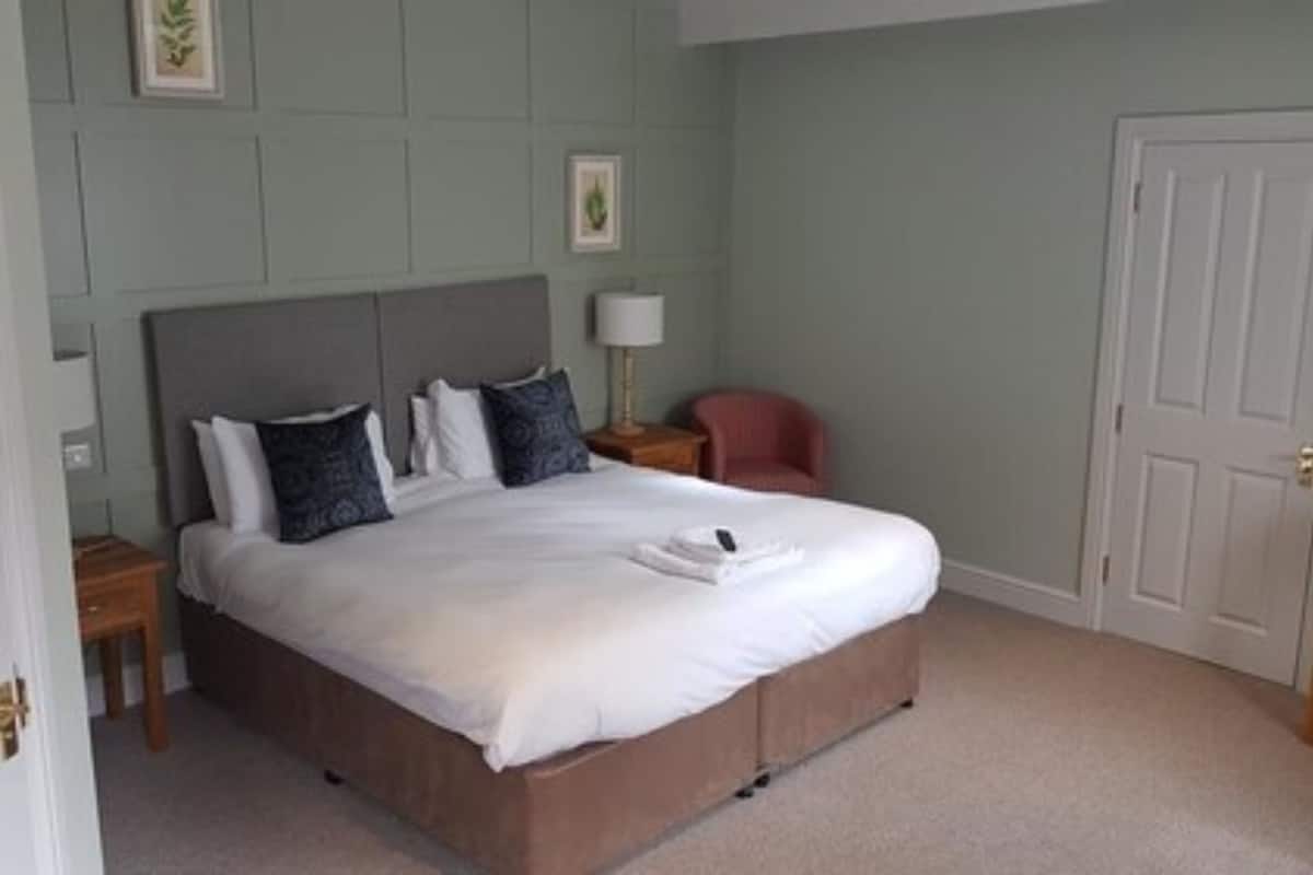 Una cama bien hecha en una habitación con pared verde claro, cabecera de madera y una mesita de noche con una lámpara.