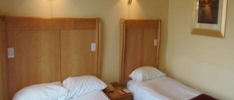 Habitación de hotel con dos camas, cada una con cabecero de madera y ropa blanca. Dos lámparas de pared a cada lado de las camas. Un cuadro enmarcado cuelga en la pared sobre las camas.